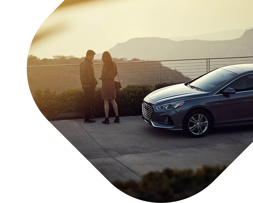 A man and a woman stand beside a sedan near a lookout point at sunset. 